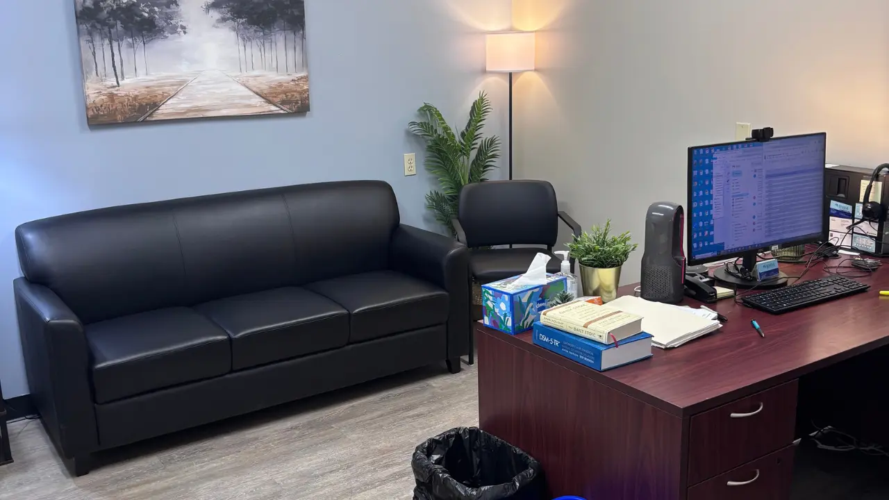 A clinic consultation office with a black leather sofa, desk, computer monitor, and medical reference books. BNBA