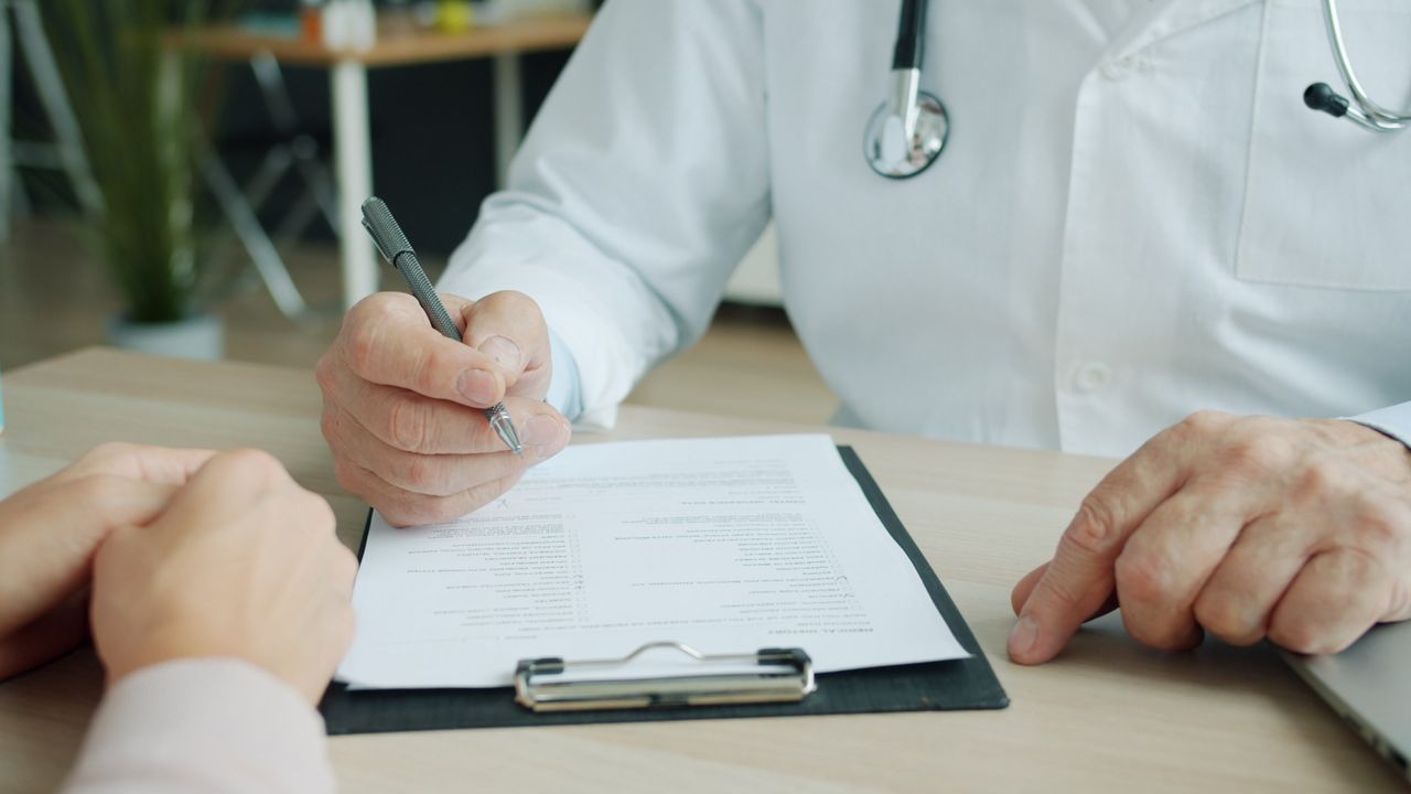 Doctor writing notes during a TMS therapy consultation with a patient across a desk. BNBA